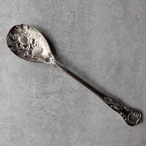 May include: Silver serving spoon with ornate floral and fruit detailing on the bowl. The handle features a decorative design with a shell-like element at the end. The spoon is set against a gray background.