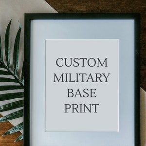 May include: A black framed print with the text "CUSTOM MILITARY BASE PRINT" on a white background. The print is sitting on a brown surface with a green plant leaf in the corner.