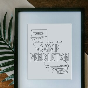 May include: A black and white line drawing of the state of California with the text "Camp Pendleton" and "Marine Corps Base" written on it. There is a helicopter flying above the state. The drawing is framed in a black frame.
