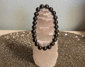 May include: A black hematite beaded bracelet on a white selenite crystal stand. The bracelet is made of round beads and is approximately 7 inches in length.