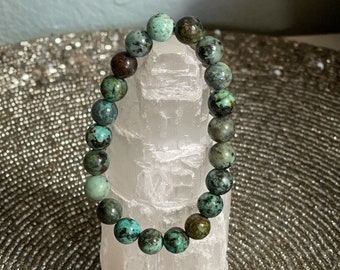 May include: A turquoise gemstone bracelet with green and brown accents. The bracelet is made of round beads and is strung on a clear elastic cord.