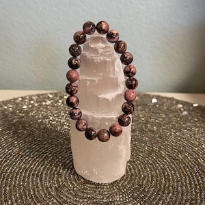 Könnte beinhalten: Ein Armband aus rosa und schwarzen Edelsteinen mit runden Perlen. Das Armband wird auf einem weißen Selenitkristall präsentiert.