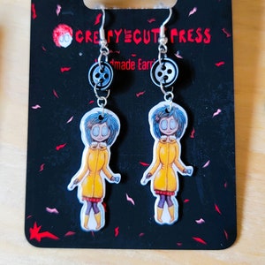 May include: A pair of handmade earrings featuring a cartoon character with blue hair and a yellow raincoat. The earrings are silver with a black button charm. The card reads "Creepycute Press Handmade Earrings".