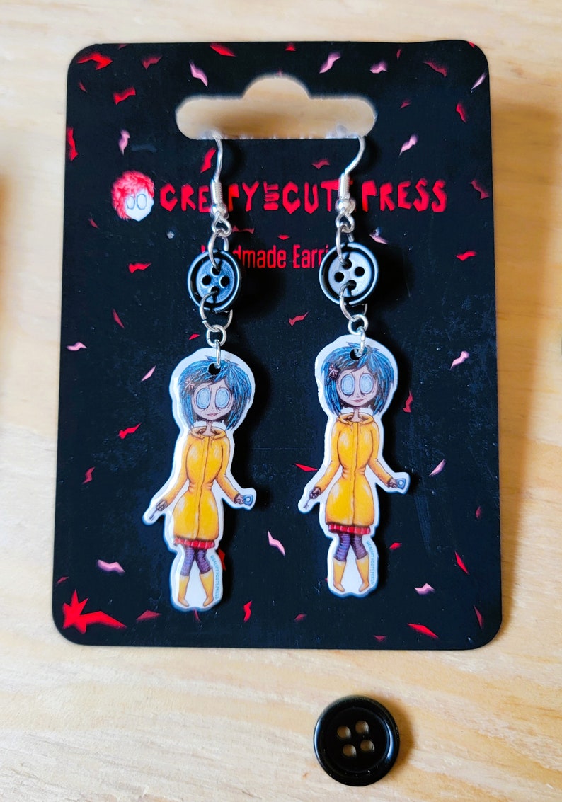 May include: A pair of handmade earrings featuring a cartoon character with blue hair and a yellow raincoat. The earrings are silver with a black button charm. The card reads "Creepycute Press Handmade Earrings".
