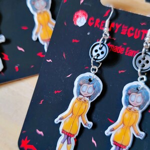 May include: A pair of earrings featuring a cartoon character with blue hair and a yellow raincoat. The earrings are made with black button accents and silver hooks. The card reads "Creepy Cute Made Earrings".