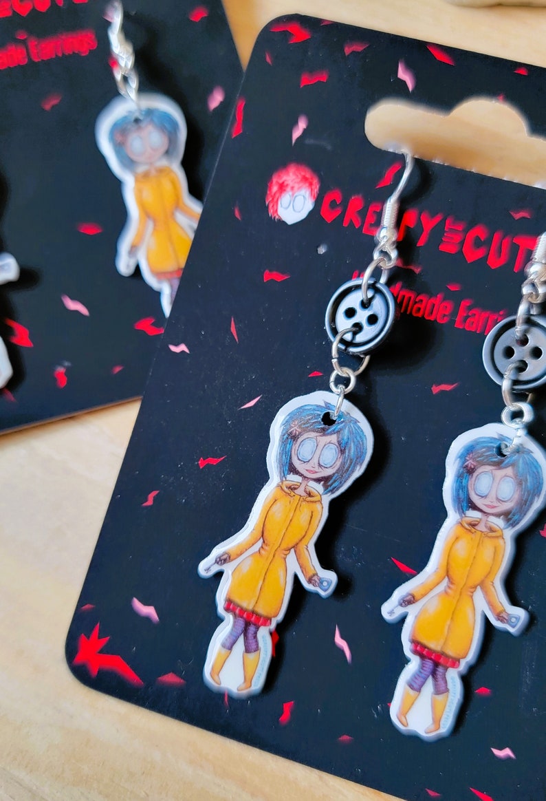 May include: A pair of earrings featuring a cartoon character with blue hair and a yellow raincoat. The earrings are made with black button accents and silver hooks. The card reads "Creepy Cute Made Earrings".