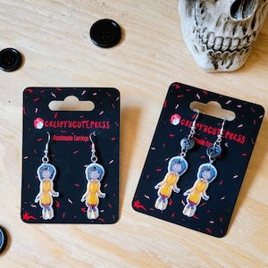 May include: A pair of handmade earrings featuring a cartoon character with blue hair wearing a yellow raincoat. The earrings are on black card stock with the text "Creepy Cute Press" and "Handmade Earrings".