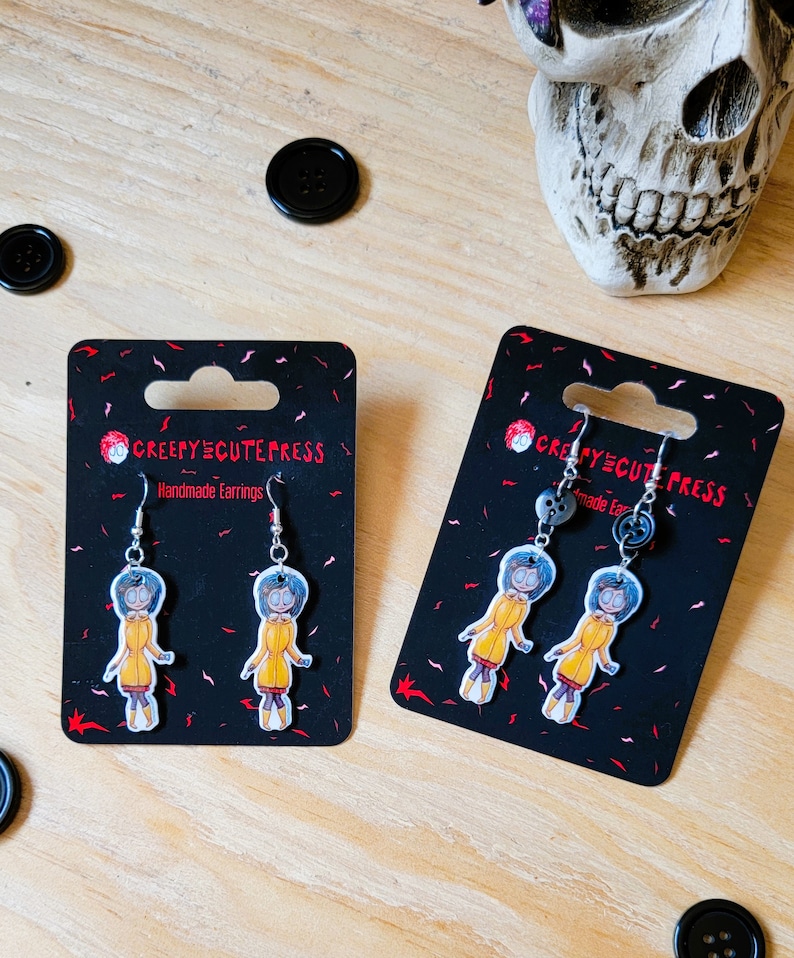 May include: A pair of handmade earrings featuring a cartoon character with blue hair wearing a yellow raincoat. The earrings are on black card stock with the text "Creepy Cute Press" and "Handmade Earrings".