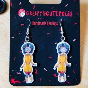May include: A pair of silver earrings with a cartoon character wearing a yellow raincoat and blue hair. The earrings are on a black card with the text "Creepy Cute Press Handmade Earrings".