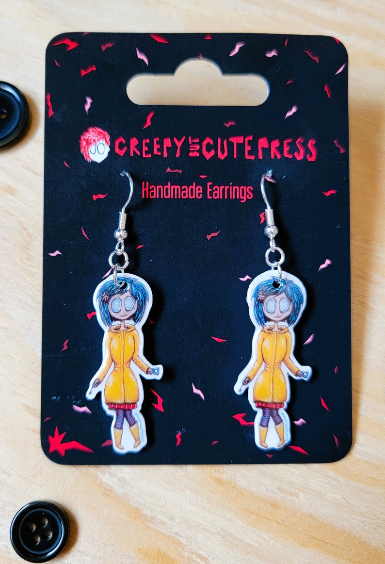 May include: A pair of silver earrings with a cartoon character wearing a yellow raincoat and blue hair. The earrings are on a black card with the text "Creepy Cute Press Handmade Earrings".