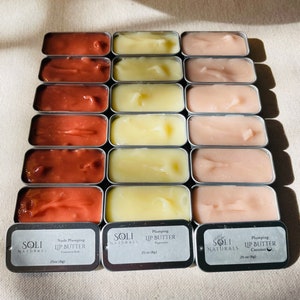 May include: A collection of 12 small, round, metal tins filled with lip butter. The tins are arranged in three rows of four. The lip butter is in various shades of pink, red, and yellow. The tins have labels that say "SoLi Naturals" and "Lip Butter".