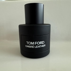 May include: A matte black cylindrical perfume bottle with a matching cap. The front features a rectangular black label with the white text TOM FORD OMBRÉ LEATHER printed in a clean, sans-serif font. The design is sleek, opaque, and uniform in color.