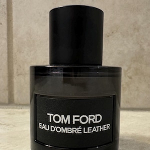 May include: A black Tom Ford Eau d'Ombré Leather fragrance bottle. The bottle is rectangular with a rounded base and a cylindrical cap. The label features the brand name and fragrance name in white text.
