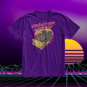 Może przedstawiać: Fioletowy T-shirt z retro grafiką buldożera z tekstem "Cruisin' for a Bruisin'" w różowych i zielonych literach.