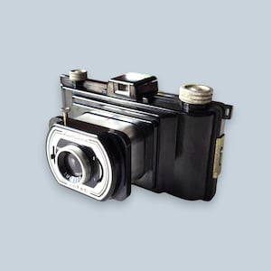 Puede incluir: Una cámara plegable vintage negra Lumière Lutac con cuerpo rectangular, lente plateada y perillas de ajuste metálicas en la parte superior. La placa frontal presenta el nombre de la marca Lumière y el modelo Lutac en letras blancas.