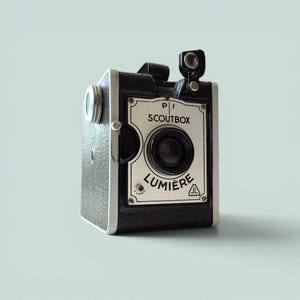 May include: A black and silver box camera with a textured finish. The front plate features the text PI SCOUTBOX and LUMIERE, along with a central lens, a small viewfinder at the top, and two small circular icons representing a flash and a tripod.