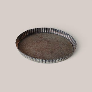May include: A round, shallow metal tart pan with a fluted edge. The surface of the pan features a dark, weathered, and oxidized finish with visible brown and grey rust-like textures throughout the interior and along the exterior fluted sides.