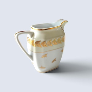 Może przedstawiać: Biały dzbanek na śmietankę z porcelany ze złoconym brzegiem i uchwytem. Górna część posiada poziomy pas ze wzorami złotych liści, natomiast dolna część korpusu jest ozdobiona małymi, rozproszonymi motywami złotych liści na gładkiej powierzchni.