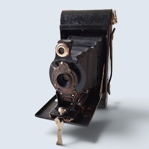 Pode incluir: Uma câmara dobrável preta vintage com mecanismo de fole, caixa de lente circular e detalhes metálicos. A placa frontal apresenta o texto "No. 2 Folding Autographic Brownie" em letras douradas, com uma pequena alavanca de metal prateado na base.