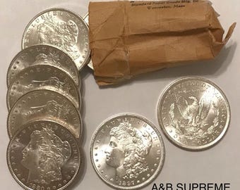 Op de afbeelding: Een verzameling zilveren Morgan dollar munten, sommige gedateerd 1880 en 1897, met gedetailleerde gravures van Lady Liberty en een adelaar. Er is ook een bruin papieren pakket, met de tekst "Standard Paper Goods Mfg. Co. Worcester, Mass."