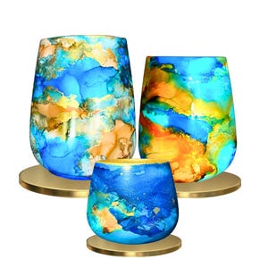 Könnte beinhalten: Drei gläserne Kerzenhalter mit abstrakter Marmorierung in Blau, Orange und Gold. Die Kerzenhalter stehen auf goldenen Metallsockeln.