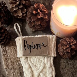 May include: A white knitted Christmas stocking with a brown leather tag that says "Bagheera" with a heart symbol. The stocking is hanging on a stone fireplace with pine cones and a candle.