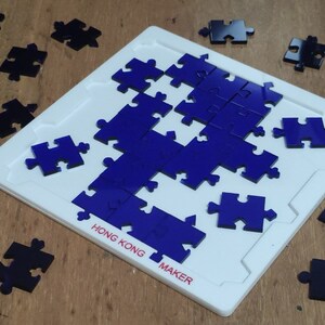 Puede incluir: Un rompecabezas de madera azul con 18 piezas en una tabla blanca. La tabla tiene el texto "HONG KONG MAKER" impreso en ella. Las piezas del rompecabezas están dispersas alrededor de la tabla.