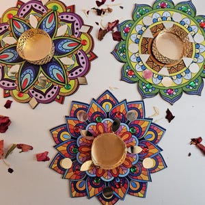 May include: Three decorative mandala candle holders. Each features a gold-coloured metal cup for a candle in the centre. The mandalas are adorned with vibrant colours, mirrored details, and gemstones. Dried rose petals are scattered around the candle holders.