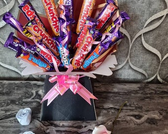 May include: A bouquet of chocolate bars arranged in a pink and black box with a pink ribbon. The chocolate bars are wrapped in purple and gold foil and include brands like Crunch, Boost, and Fudge.