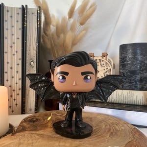 Op de afbeelding: Een vinyl verzamelfiguur van een personage met donker haar, een lichte huid en paarse oogschaduw. Het figuurtje draagt een zwart pak en heeft grote, getextureerde zwarte vleermuisvleugels op de rug. Het staat op een ronde zwarte voet.