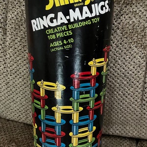 May include: A Slinky brand Ringa-Majigs toy in a black and red cylindrical container. The container has a picture of the toy on it, which is a stack of colorful rings and poles. The text on the container reads "Slinky Brand Ringa-Majigs Creative Building Toy 108 Pieces Ages 4-10 (Actual Size)"