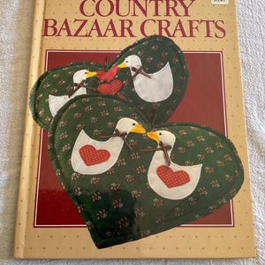 May include: A book titled "Better Homes and Gardens Country Bazaar Crafts" with heart-shaped quilted potholders featuring duck and heart designs. The potholders are green with white ducks, red hearts, and yellow beaks.