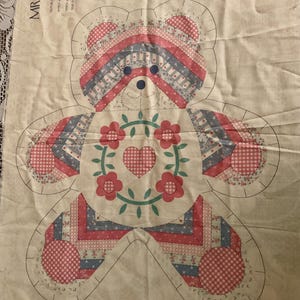 May include: A cream-colored fabric panel featuring a printed pattern for a stuffed bear named MR. TED E. BEAR. The design includes a bear outline with red, blue, and white gingham and floral patterns, a central heart surrounded by flowers, and text.
