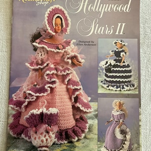 May include: A crochet pattern book titled "Hollywood Stars II" featuring doll designs. The cover showcases a doll in a pink and purple ruffled gown and bonnet. Additional doll designs are shown in the lower right and upper right corners.