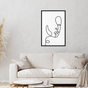 May include: A black and white line drawing of a face in a black frame hanging on a white wall above a white couch with two gold nesting tables.