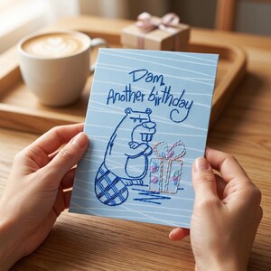 Puede incluir: Tarjeta de cumpleaños azul claro con una ilustración de castor y el texto "Dam, Another birthday". El castor sostiene una caja de regalo. La tarjeta se sostiene frente a una taza de café y una pequeña caja de regalo.