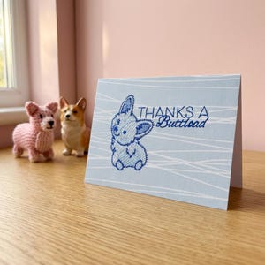 May include: A light blue greeting card with a stitched bunny design and the text "THANKS A Buttload". Two corgi dog figurines are in the background, one pink knitted and one ceramic.