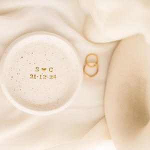May include: A small, speckled, off-white ceramic ring dish with gold lettering and a heart symbol. The dish displays the initials "S C" and the date "21-12-24". Two gold rings rest beside the dish on a cream-colored fabric.