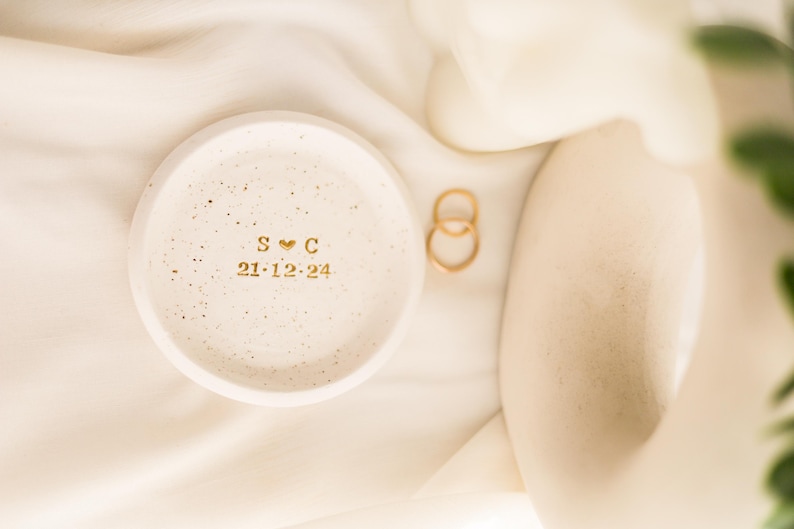 May include: A small, speckled, off-white ceramic ring dish with gold lettering and a heart symbol. The dish displays the initials "S C" and the date "21-12-24". Two gold rings rest beside the dish on a cream-colored fabric.