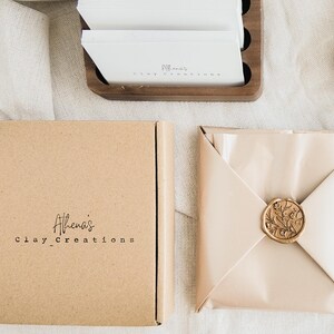 May include: A square, light brown cardboard box with the text "Athena's clay_Creations" in black. Beside it, a package wrapped in light beige paper, sealed with a gold wax stamp. A wooden holder with white cards is in the background.
