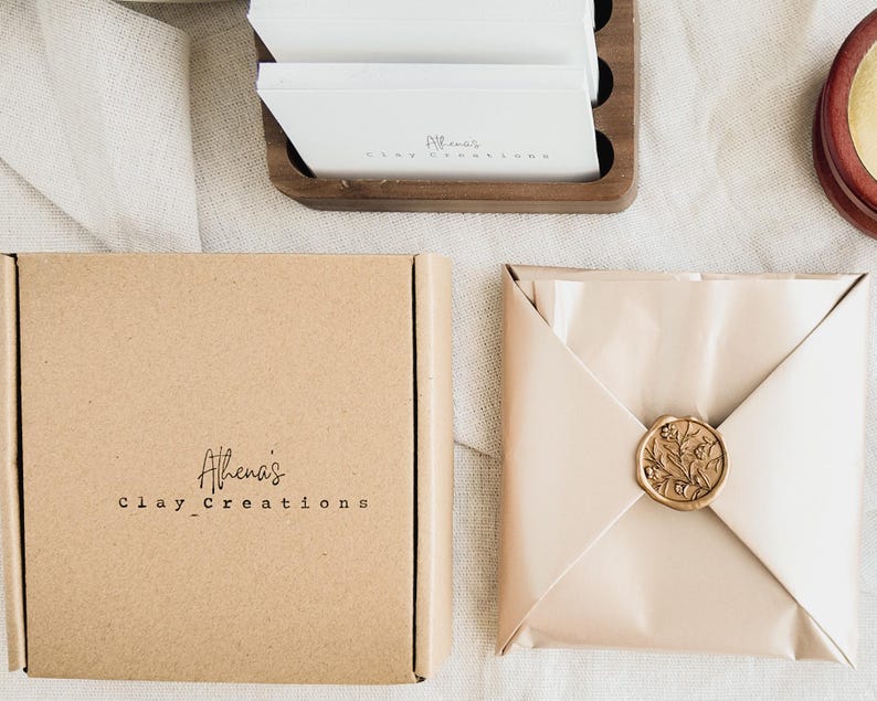May include: A square, light brown cardboard box with the text "Athena's clay_Creations" in black. Beside it, a package wrapped in light beige paper, sealed with a gold wax stamp. A wooden holder with white cards is in the background.