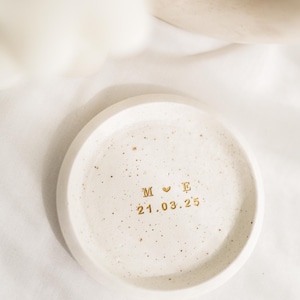 May include: A small, round, off-white ceramic dish with a speckled surface. Gold lettering includes initials "M E" with a heart symbol and the date "21.03.25". The dish is likely used for holding small items.