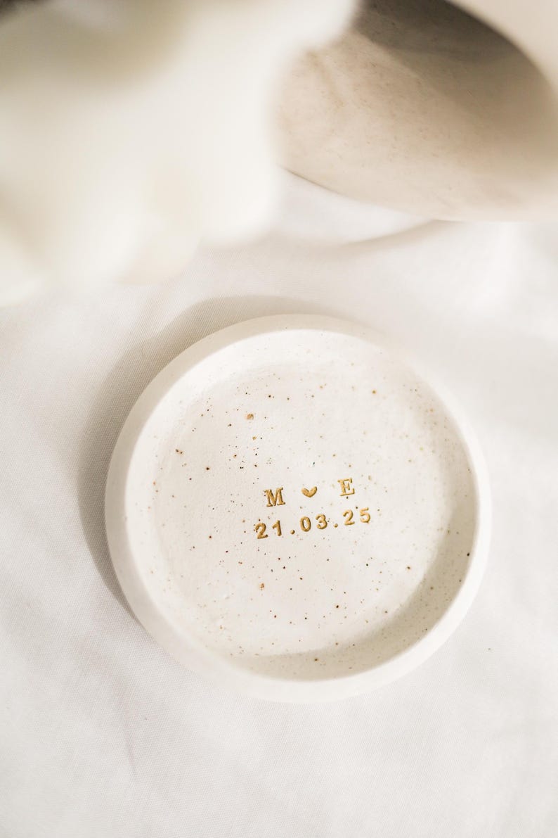 May include: A small, round, off-white ceramic dish with a speckled surface. Gold lettering includes initials "M E" with a heart symbol and the date "21.03.25". The dish is likely used for holding small items.