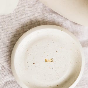 May include: A speckled, off-white ceramic dish with a slightly raised edge. The word "Madi" is written in gold lettering in the centre. The dish is on a light-coloured, textured surface.