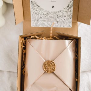 May include: A gift box with a beige-colored wrapped package sealed with a gold wax seal. The seal features a floral design. A card with the word "Congratulations" and the name "Danni" is visible inside the box.
