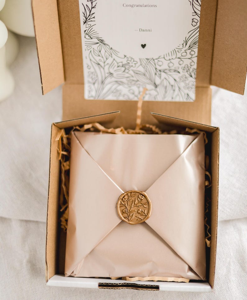 May include: A gift box with a beige-colored wrapped package sealed with a gold wax seal. The seal features a floral design. A card with the word "Congratulations" and the name "Danni" is visible inside the box.