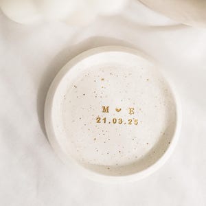 May include: A small, round, speckled, off-white ceramic tray. The tray has gold lettering that reads "M ♡ E" above the date "21.03.25". The tray is likely used for holding small items like jewelry.