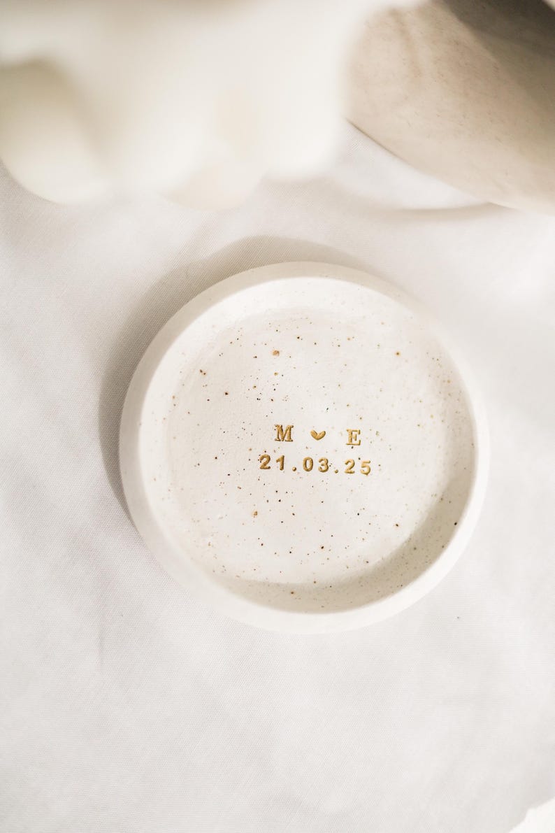 May include: A small, round, speckled, off-white ceramic tray. The tray has gold lettering that reads "M ♡ E" above the date "21.03.25". The tray is likely used for holding small items like jewelry.