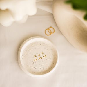 May include: A small, round, speckled ceramic tray with gold lettering. The tray has the initials "E C" and the date "14.02.25" in gold. Two gold rings are next to the tray. The background is a white cloth.