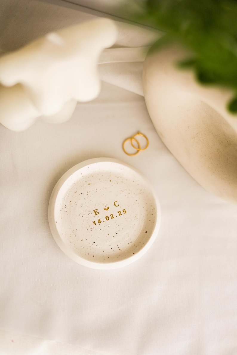 May include: A small, round, speckled ceramic tray with gold lettering. The tray has the initials "E C" and the date "14.02.25" in gold. Two gold rings are next to the tray. The background is a white cloth.